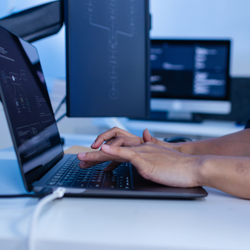 Close-up of hands typing on a laptop keyboard with multiple monitors displaying code in a dark blue-lit workspace