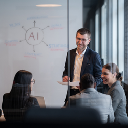 A smiling man in a blazer leads a meeting with colleagues at a table, with an AI diagram drawn on a whiteboard behind him