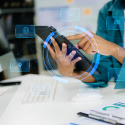 Person holding a smartphone with a glowing circular digital interface and icons overlaid, at a desk with charts and a keyboard