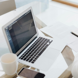 Overhead view of an open laptop on a glass desk next to a coffee mug, notebook, and pen in a bright minimal workspace