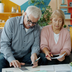 An older man and woman sitting together on a gray couch in a brightly colored living room. The man, wearing glasses and a plaid shirt, is using a smartphone and holding some cash. The woman, wearing a pink sweater, holds a pen and looks at documents spread out on a coffee table. They look focused on reviewing their paperwork and finances.