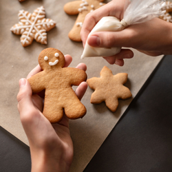 Two hands decorate a gingerbread man cookie with white icing using a small plastic piping bag. The left hand holds the cookie, which has just received two dots for eyes and a small curved smile, while the right hand squeezes the icing bag directly above it. The scene is set over a sheet of brown parchment paper on a dark surface, surrounded by other baked holiday cookies, including a heavily decorated snowflake and plain star-shaped cutouts in the background.