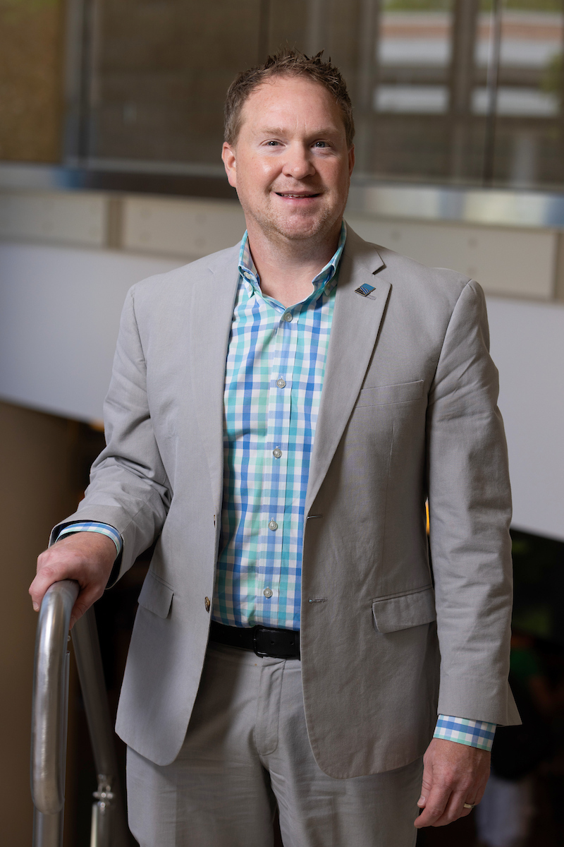 Person wearing a light gray suit jacket and checked button-down shirt, standing indoors with one hand resting on a railing.