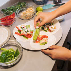 Alt text: "Hands assembling tacos on white tortillas, topped with shrimp, sliced red peppers, avocado, and fresh herbs, surrounded by bowls of colorful ingredients on a counter.