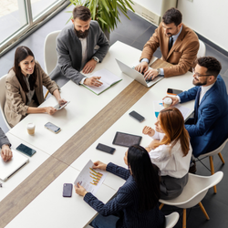 High-angle view of a group of business professionals sitting around a long conference table, collaborating with laptops, tablets, and printed reports.