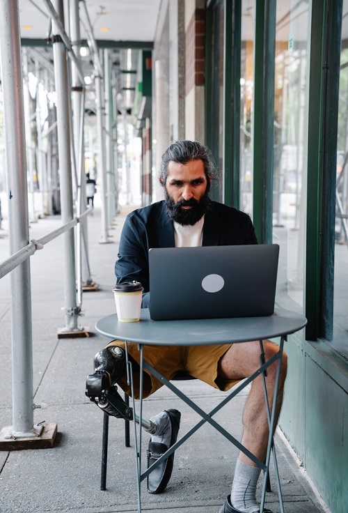Man with prosthetic leg siting at an outdoor table with laptop and coffee