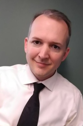 Stephen Kelly smiles at the camera wearing a white button up with a black tie