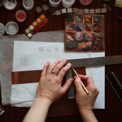 Hands of a jeweler measuring a copper strip on a cluttered workbench with enamel paints and metalwork samples.