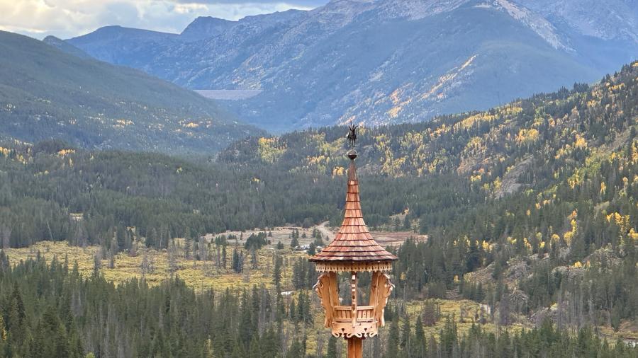 A wooden tower structure with a carved roof set against a dramatic mountain valley landscape with layers of forest and peaks.