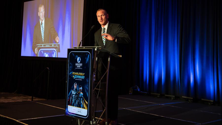 A person in a suit stands at a podium on a stage, delivering a speech at an event with blue stage lighting and a large screen displaying the same podium scene behind them.