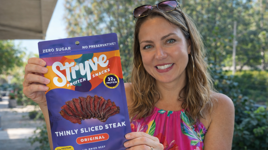Person holding a colorful package of Stryve protein snacks labeled ‘Thinly Sliced Steak – Original’ while standing outdoors on a sunny day.