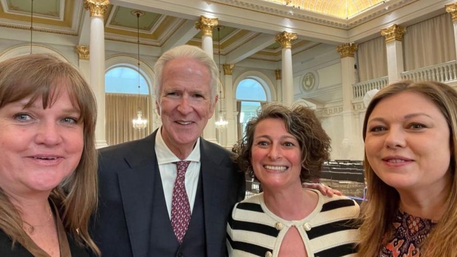 Four people stand closely together in an ornate hall with high ceilings, white columns, gold architectural details, and large chandeliers. They are dressed in formal or semi-formal attire and are posing for a group photo in front of rows of empty seats.