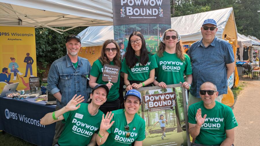 A group of people wearing green “Powwow Bound” shirts poses in front of a large “Powwow Bound” banner at an outdoor event, surrounded by tents and PBS Wisconsin displays. 