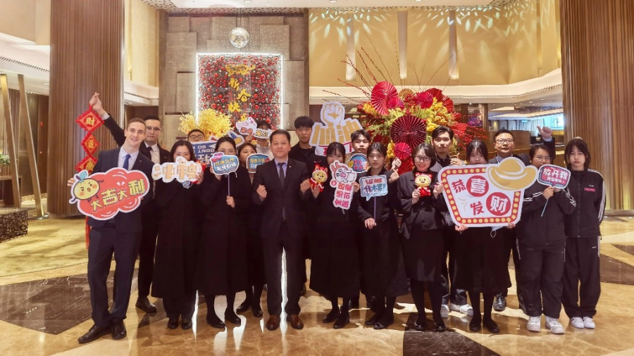 A large group of people stands together in the lobby of an upscale building with tall columns, warm lighting, and decorative floral displays featuring red and gold colors. The group holds festive signs with cartoon-style graphics and bright colors, including icons resembling flowers, hats, and celebratory symbols. The setting includes polished floors, a high ceiling with curved light fixtures, and a large, ornate floral arrangement on the back wall.