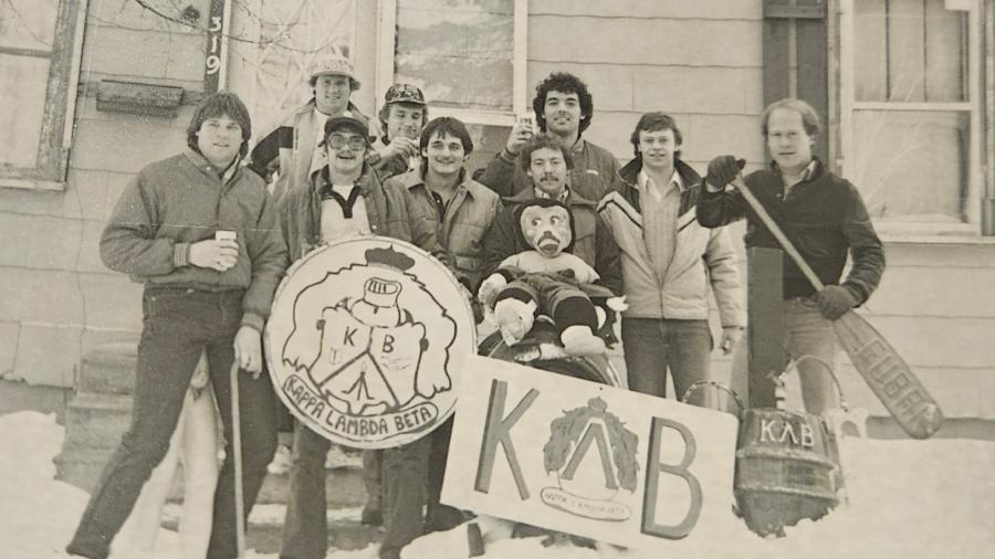 A group of people stands outdoors in the snow in front of a house, holding large KLB-themed signs and props. Several individuals wear winter jackets, and one person holds a paddle marked with “KΛB.” Snow covers the ground and the house exterior is visible behind them.