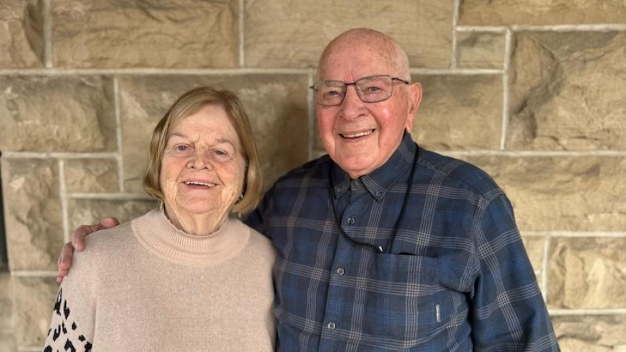 Two older adults standing side by side in front of a stone wall, with one person’s arm around the other, both facing the camera.