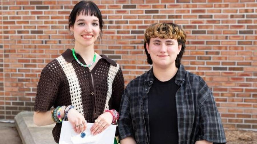 Two people stand side by side outdoors in front of a brick wall. One holds an envelope labeled “Stout University Foundation Scholarship.”