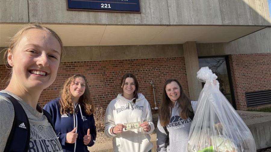 UW-Stout tennis team volunteers together outdoors, posing near a campus building while holding a large bag of collected trash.