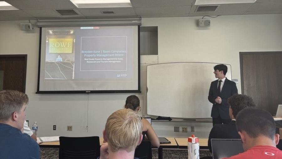 A person in a suit stands at the front of a classroom presenting slides to seated attendees, with a projector screen and whiteboard visible.
