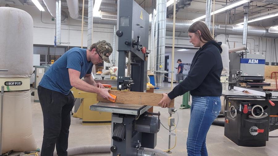 Konnor Kraft cuts pieces for a ukulele neck on a bandsaw, assisted by TA Annika Schleuter