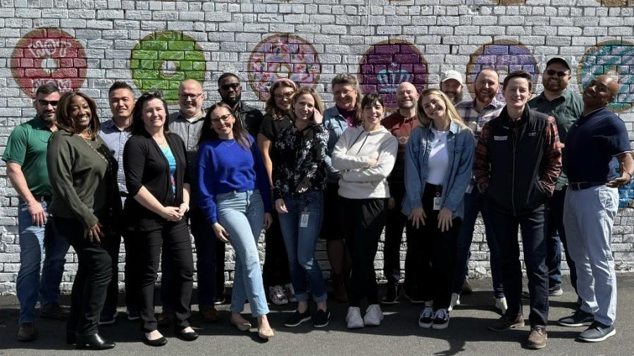 Group of about 15 people standing outdoors in front of a brick wall painted with colorful donut designs, posing closely together for a photo.