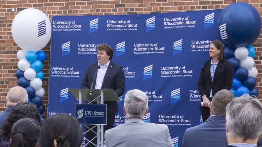 Speaker stands at a podium during a UW-Stout groundbreaking ceremony, with a branded backdrop and audience in front.