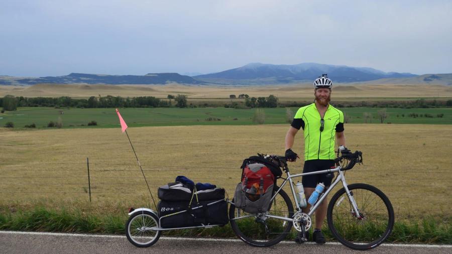 Person in a bright cycling jersey stands beside a fully loaded touring bicycle on a rural roadside, with open fields and distant mountains in the background.