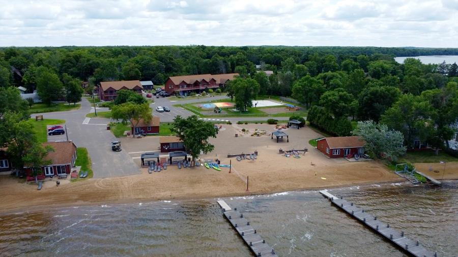 Aerial view of a lakeside resort featuring sandy beach, docks, cabins, and a wooded shoreline surrounding the property.