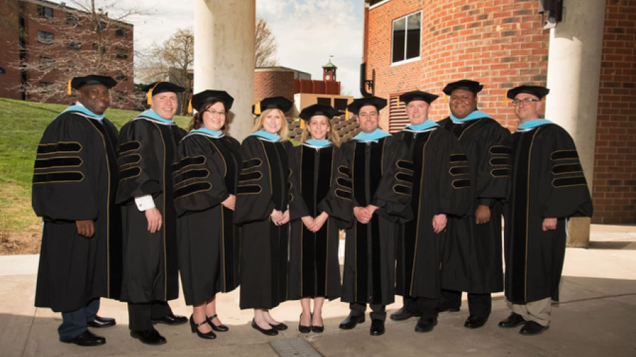 A group of students wearing black academic regalia with gold trim and caps stand outdoors on a campus walkway near brick buildings.