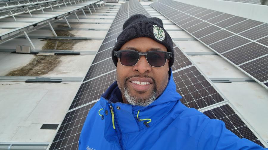 Person wearing a blue jacket and black knit hat stands on a rooftop surrounded by rows of solar panels under a cloudy sky.