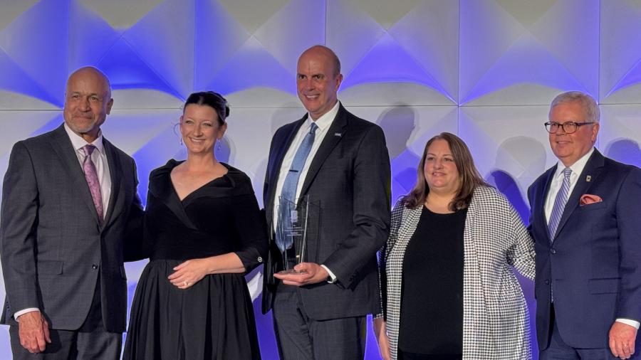 Five people pose onstage at an awards ceremony. One person in a dark suit holds a glass award while standing in front of a geometric wall illuminated with blue and purple lighting.
