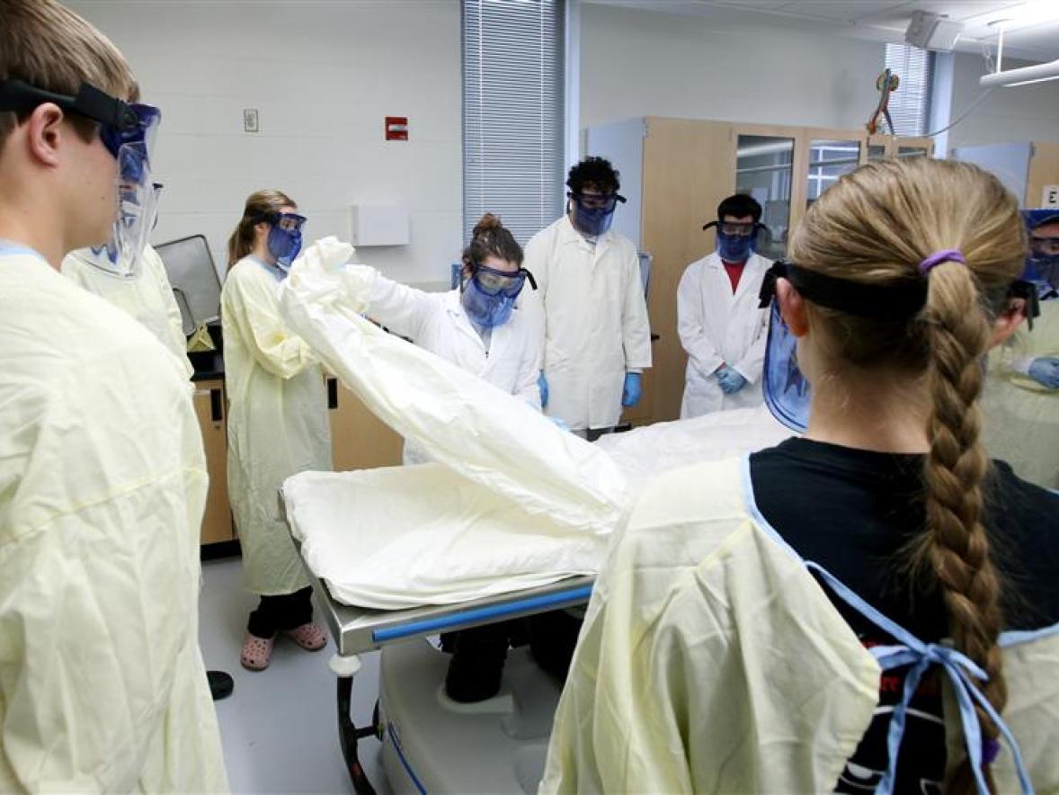 A group of students in white coats stand around a dissection table