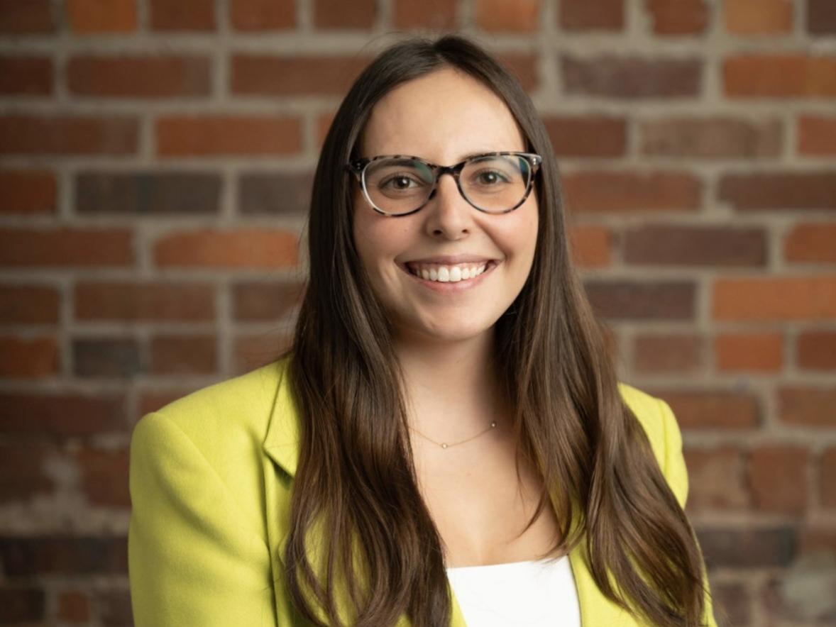 Professional headshot of a person with long dark hair wearing glasses, a bright green blazer, and a white top, standing in front of a brick wall and facing the camera.