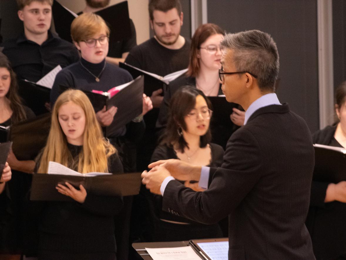A choir in formal attire performing, conducted by Dr. Jerry Hui standing in front