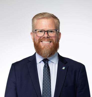 Nic Katona is shown smiling. He is wearing a navy suit along with a button down shirt and tie.