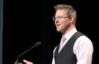 Nels Paulson, a white man with blonde hair wearing a white shirt and a vest, speaking at a podium