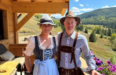 Two people wearing traditional Bavarian-style clothing standing on a wooden deck with views of rolling hills, wildflowers, and a sunny mountain landscape.