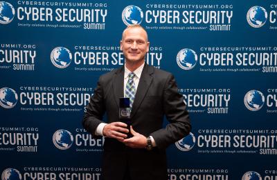 A person in a suit stands in front of a Cyber Security Summit step‑and‑repeat backdrop while holding an award.