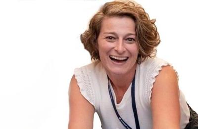 A person is leaning forward over a large inflatable globe, resting their hands on it while smiling toward the camera. They are wearing a sleeveless white top and a lanyard, with a watch on one wrist. The background is plain white.