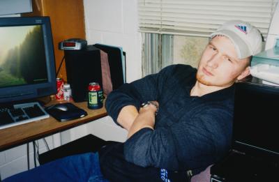 A person sits in a college dorm room with arms crossed, next to a computer desk that holds a monitor, keyboard, speakers, and soda cans.