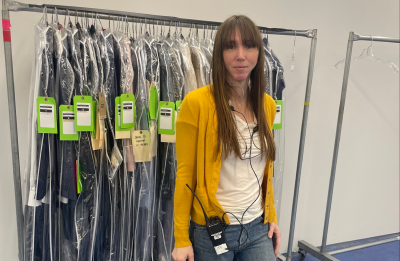 A person stands in a wardrobe workspace holding a transparent set bag. They are wearing a yellow cardigan, white shirt, and patchwork denim pants with sneakers. Behind them is a rolling rack filled with neatly organized costumes in garment bags, each tagged with bright green labels. The setting appears to be a production or costume department area with blue flooring and additional empty clothing racks nearby.