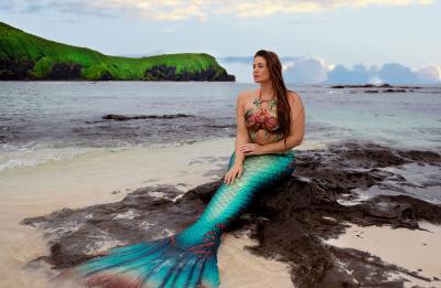 A person wearing a mermaid tail sits on rocks on a sandy beach in front of the ocean and a blue sky.