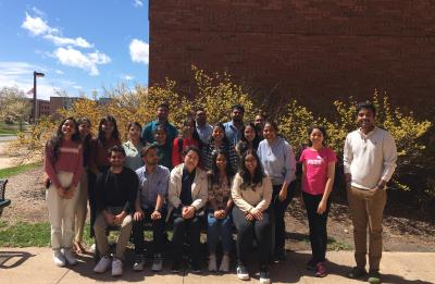 A large group of people poses for a photo outdoors on a sunny day. They are standing and sitting in several rows near a brick building, with blooming shrubs behind them and a clear blue sky overhead.