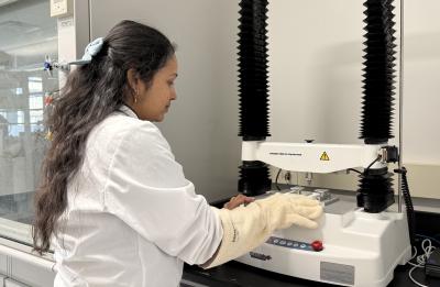A person wearing a lab coat and thick protective gloves operates a laboratory texture analyzer machine inside a lab space. Shelving, equipment, and safety features of the lab are visible in the background.
