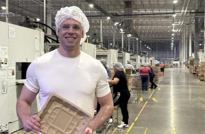 A person wearing a white T-shirt and a hairnet stands in a brightly lit industrial facility, holding a molded fiber food tray. The factory floor extends into the distance with machinery, workstations, and other workers assembling or inspecting products.