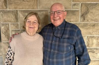 Two older adults standing side by side in front of a stone wall, with one person’s arm around the other, both facing the camera.