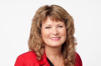 A person with shoulder‑length, wavy hair wearing a bright red blazer over a dark top, photographed against a plain white background.