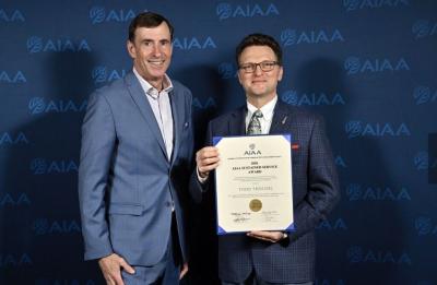 Two people stand in front of a blue AIAA-branded backdrop. One person holds a framed certificate that reads “AIAA Sustained Service Award” and includes the name “Todd Treichel.”