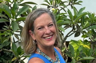Person standing outdoors in front of leafy green plants, wearing a sleeveless blue top and a beaded necklace, with a white wall partially visible in the background.