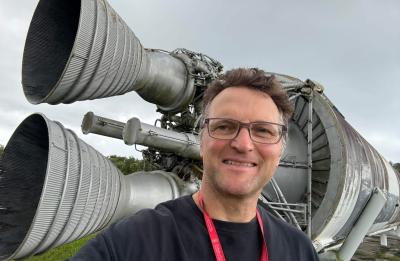 A person takes a selfie outdoors in front of two large rocket engines mounted horizontally, showing detailed metal nozzles, piping, and mechanical components against an overcast sky.